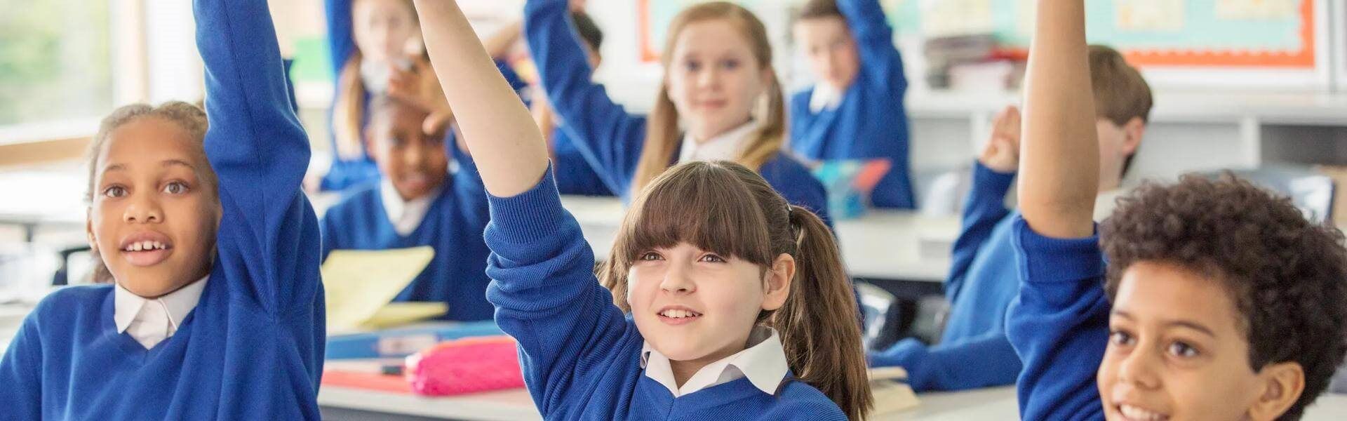 School children wearing blue school uniforms raising hands in classroom